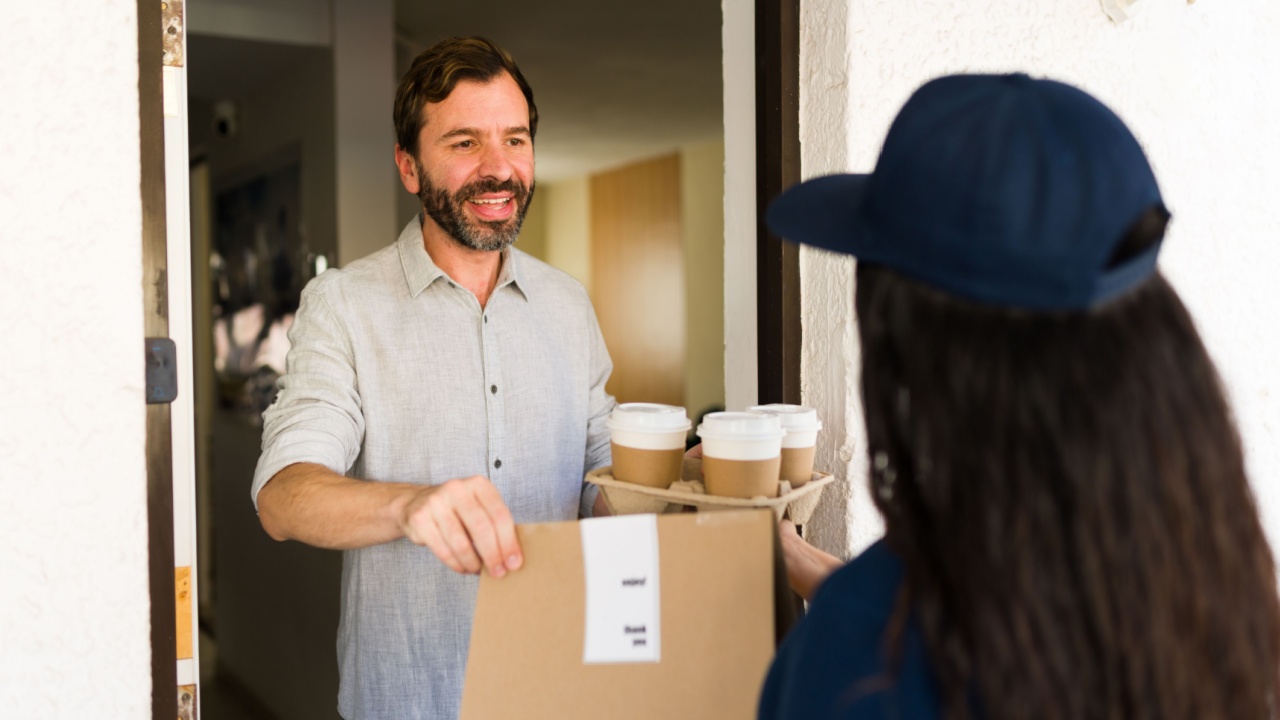 Man smiling, receiving a package and coffee cups from a delivery driver carrying a delivery box and a tray of beverages at his home front door