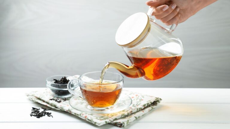 Woman pouring hot tea into cup from teapot at white wooden table,