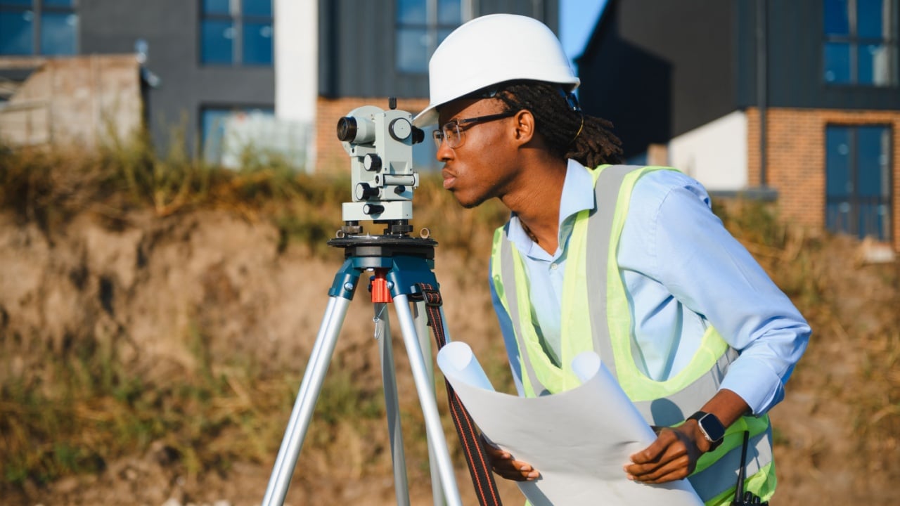 Surveyor using a theodolite for land measurement and precise leveling, holding blueprints on a new residential construction site