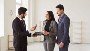 Young happy smiling couple reaching agreement with man realtor and looking through the contract of real estate sale and going to sign a contract. Real estate market and buying a property concept.