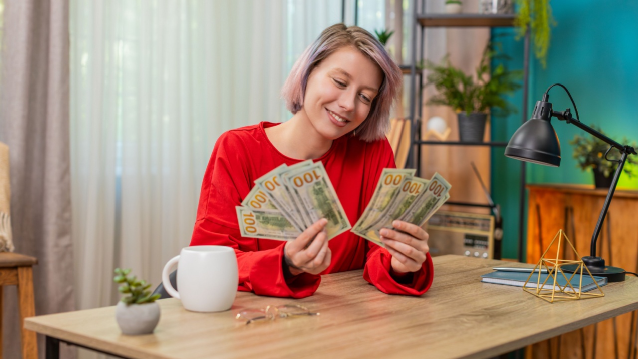 Young woman counting dollars cash money at home table, widely smiling nodding satisfied, expressing wealth. Caucasian girl cheerfully demonstrating joyful financial success and prosperity achievement.