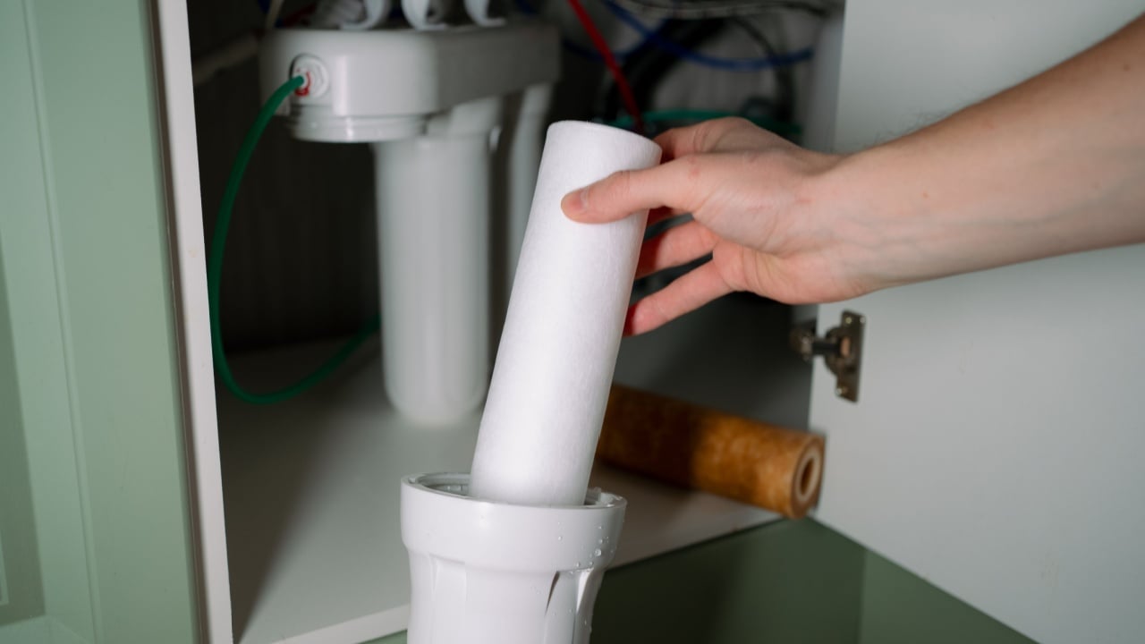 Hand installing a new water filter cartridge into an under-sink reverse osmosis unit, replacing a dirty old cartridge to restore clean drinking water for a healthy home environment