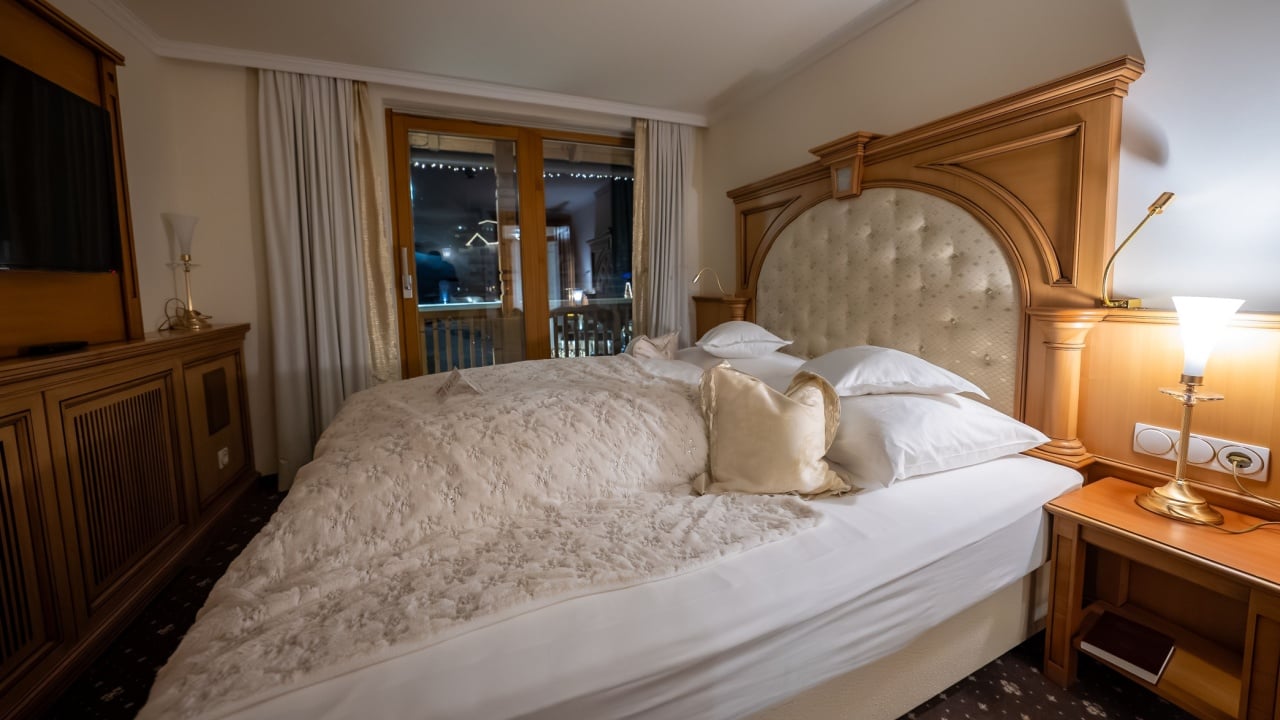Wood paneled hotel bedroom shows a large bed with tufted headboard, white linens, and a warm lamp. Glass doors reveal winter night lights and icicle like eaves in an alpine setting.
