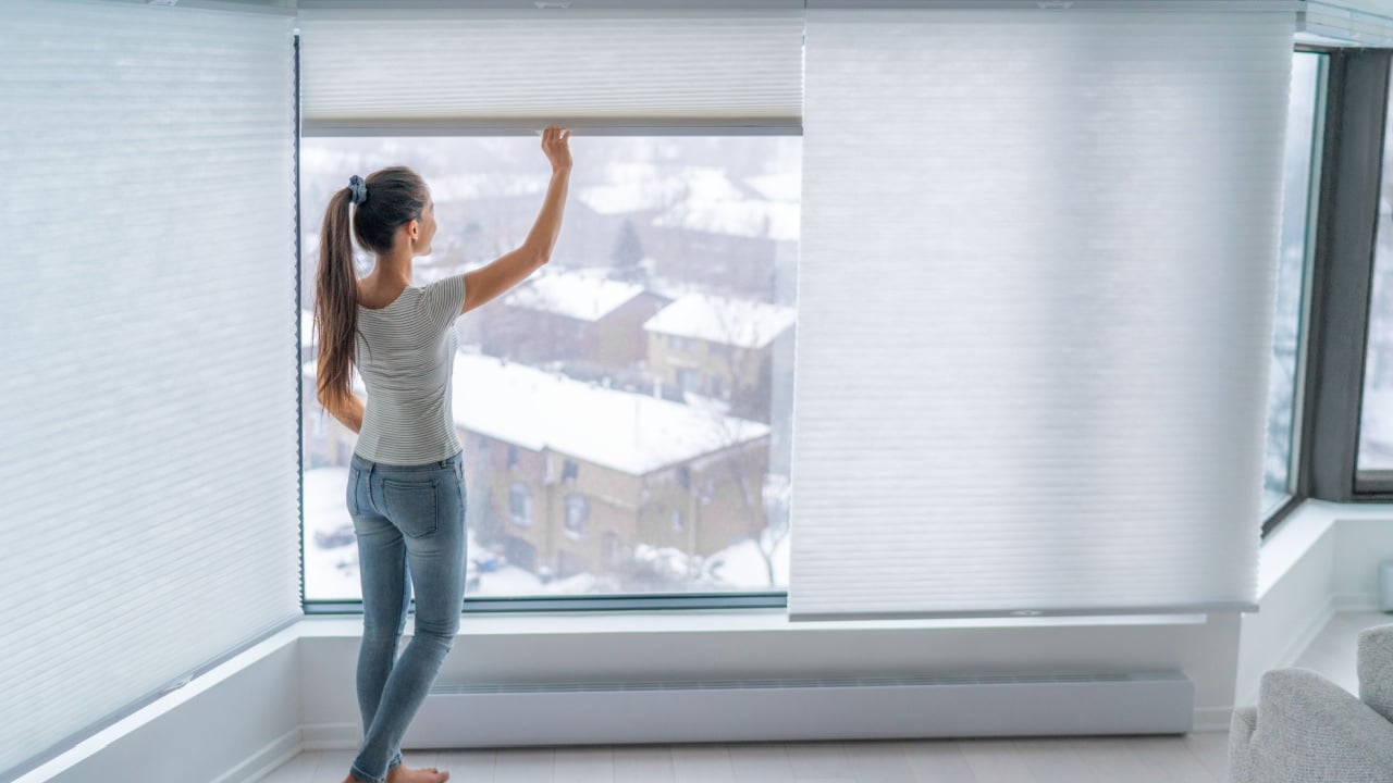 Woman closing cellular shades on apartment window keeping energy and heat indoors with honeycomb blind curtain. Cordless pleated shades in modern home living lifestyle. Interior decor design.