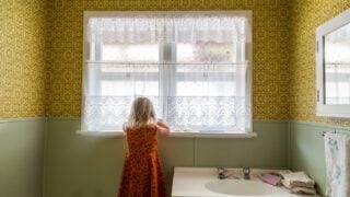 Young girl alone exploring the bathroom of a 1970s house. Lace curtains and mustard yellow colored wallpaper with green. Basin with taps and flannels. Child thinks noone is watching. Naughty.
