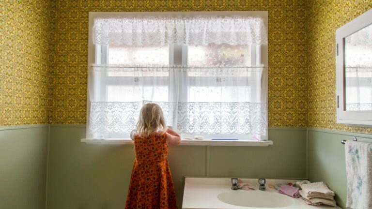 Young girl alone exploring the bathroom of a 1970s house. Lace curtains and mustard yellow colored wallpaper with green. Basin with taps and flannels. Child thinks noone is watching. Naughty.