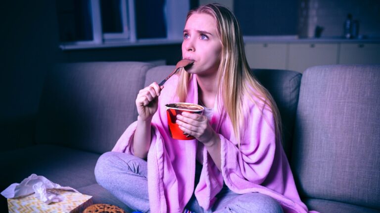Young woman watching movie at night. Concentrated model eating ice cream with spoon. Sitting on sofa in room alone. Streaming show or tv series.