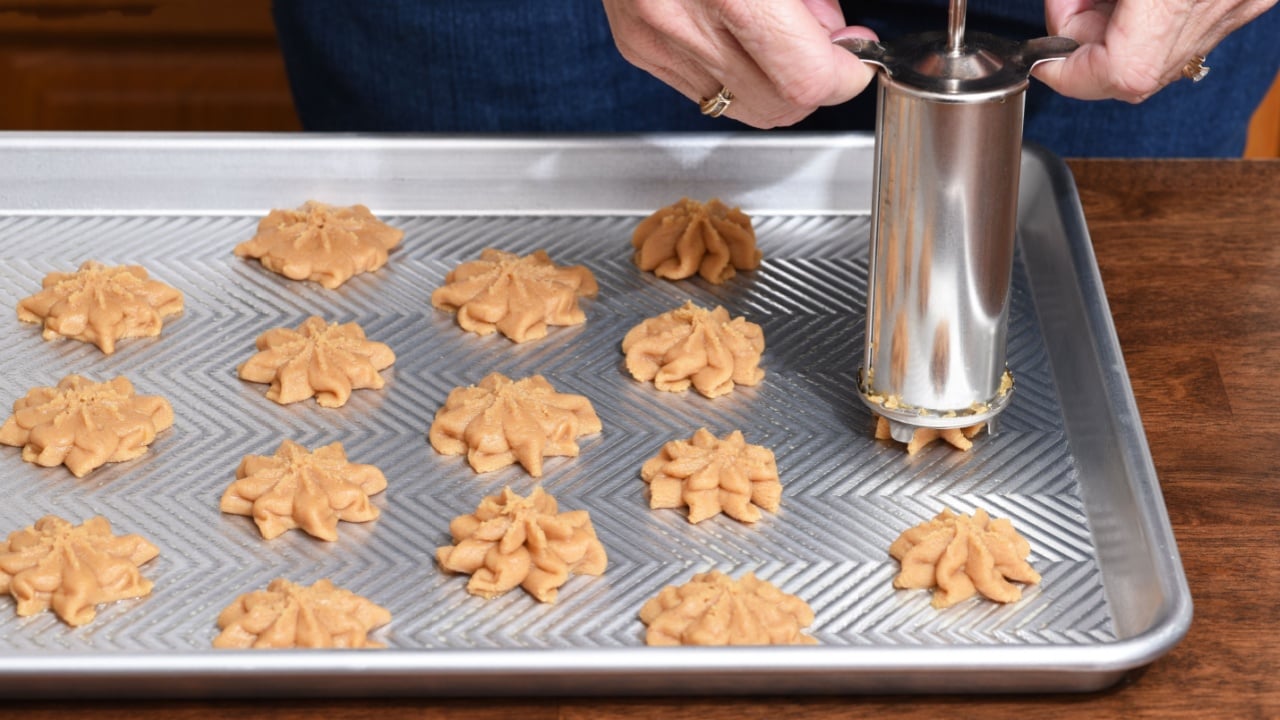 Closeup of a home baker using a cookie press to form cookies on a baking sheet. 