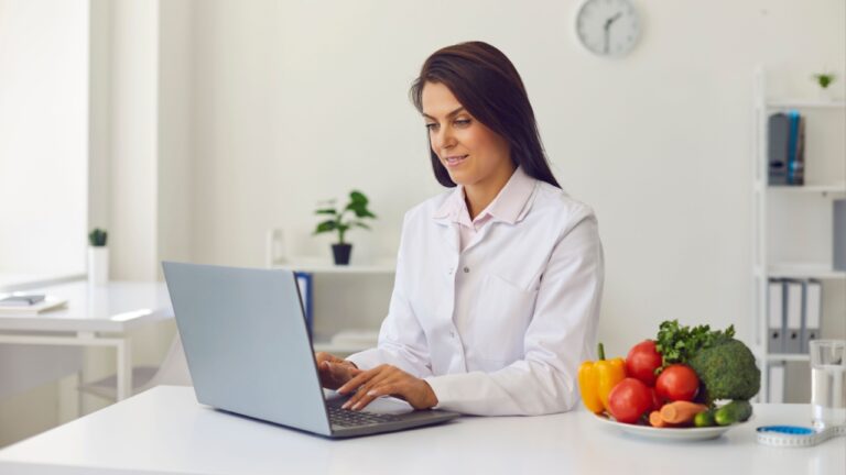 Young dietitian typing on laptop computer keyboard making balanced corrective diet plan for patient or giving detox and weight loss advice for clients online sitting at office desk with raw vegetables