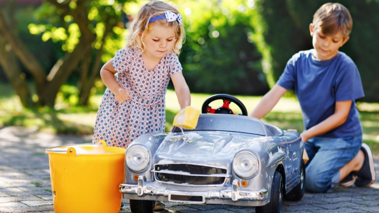 Two happy children washing big old toy car in summer garden, outdoors. Brother boy and little sister toddler girl cleaning car with soap and water, having fun with splashing and playing with sponge