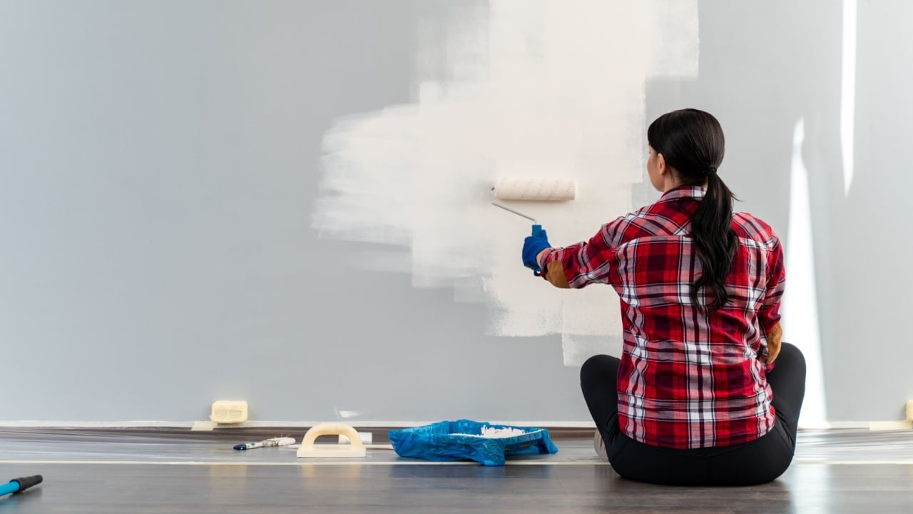 Back view of the woman sitting at the floor and painting walls with roller equipment while moving in apartment. Renovating new house and improvement concept