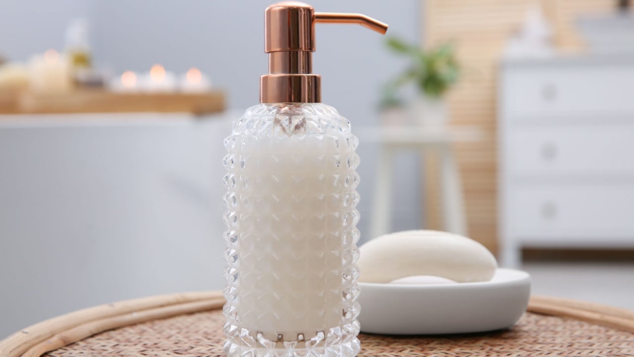 Stylish glass dispenser and soap bar on wicker stool in bathroom