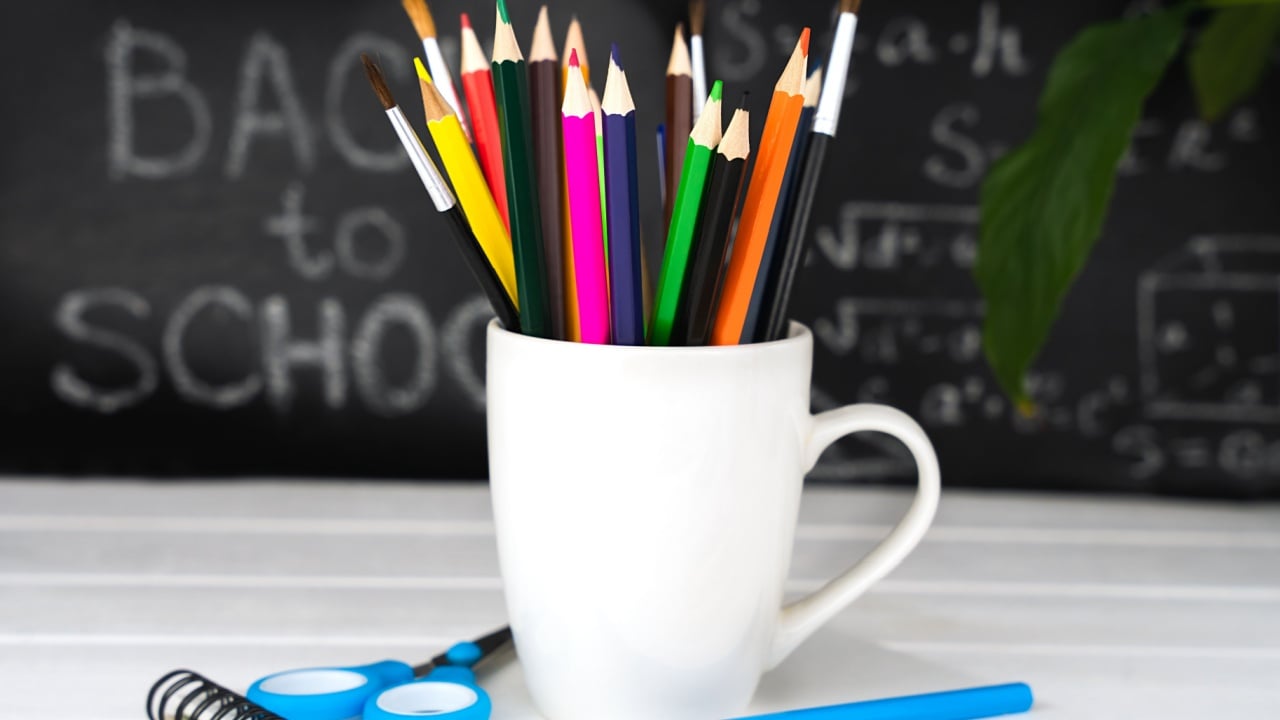 Colorful school stationery in a white mug on a wooden background. Blurred in the background is a blackboard with text. The concept of education. Selective focus.