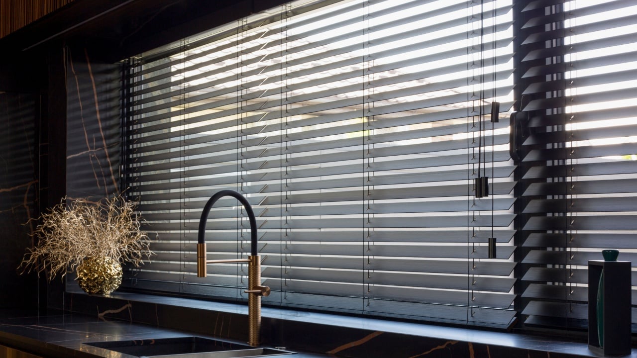 Wooden blinds black color closeup on the window. Bamboo slats 50mm wide. Venetian wood blinds in the kitchen. Black tapes. Sink with copper faucet near the window. Round vase is on the windowsill.
