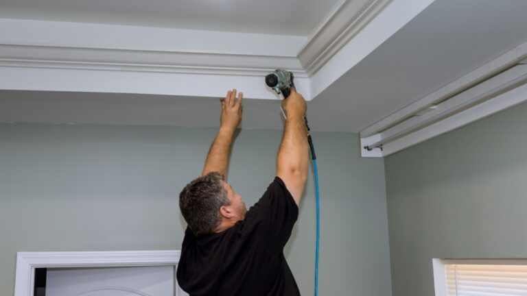 A professional carpenter nails wood crown molding between newly constructed home ceiling using an air pneumatic nail gun