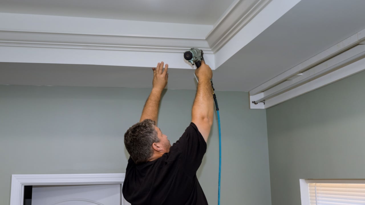 A professional carpenter nails wood crown molding between newly constructed home ceiling using an air pneumatic nail gun
