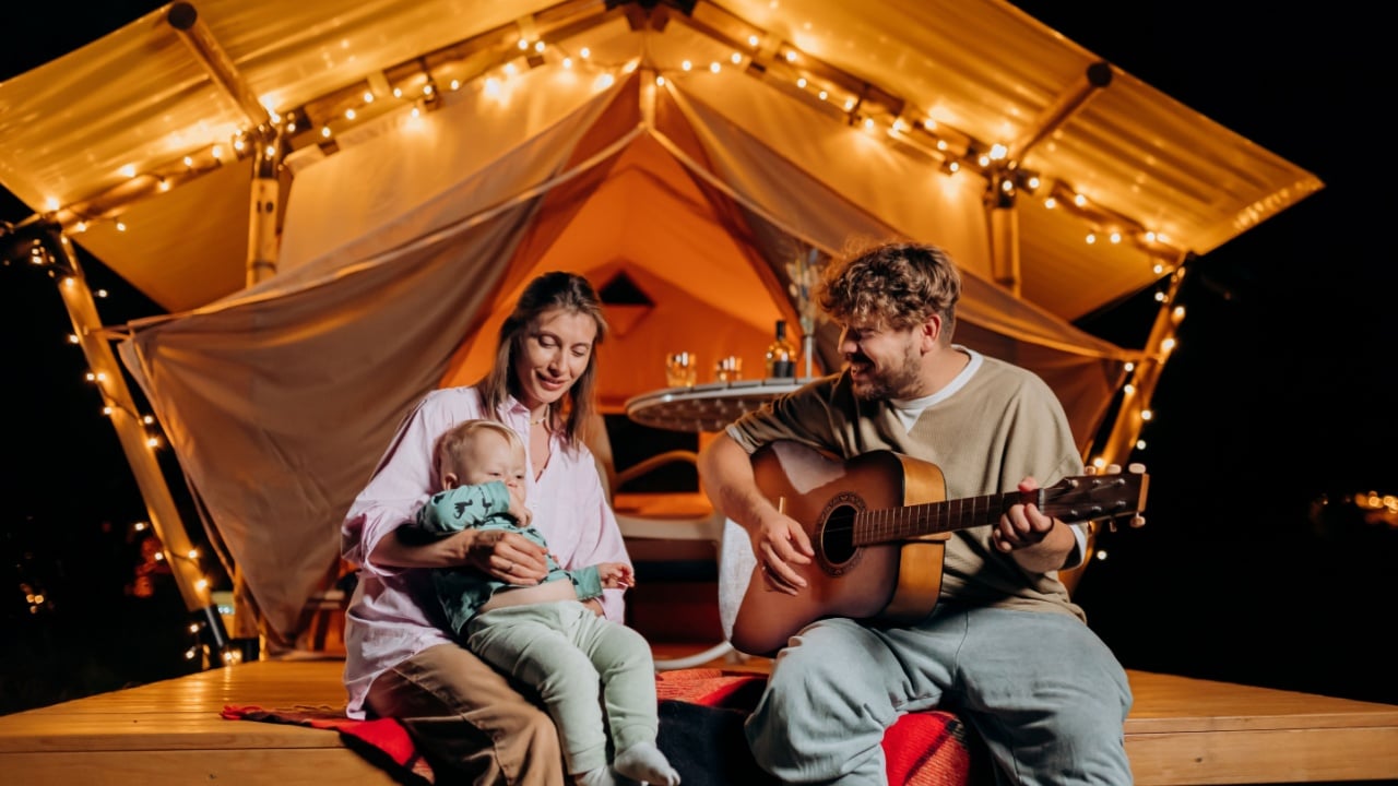 Happy family relaxing and spend time together in glamping on summer evening and playing guitar near cozy bonfire. Luxury camping tent for outdoor recreation and recreation. Lifestyle concept