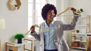 Happy woman holding empty wooden clothes hangers. Young Afro American girl in suit jacket wants to try on different sizes and fashion styles before she chooses one outfit between two on wood hangers