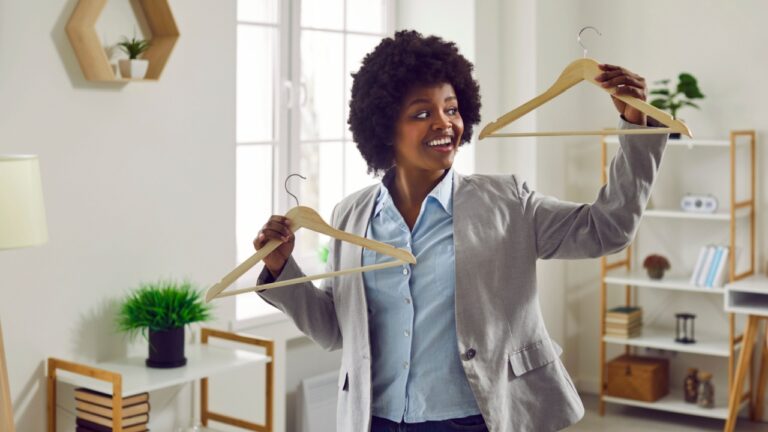 Happy woman holding empty wooden clothes hangers. Young Afro American girl in suit jacket wants to try on different sizes and fashion styles before she chooses one outfit between two on wood hangers