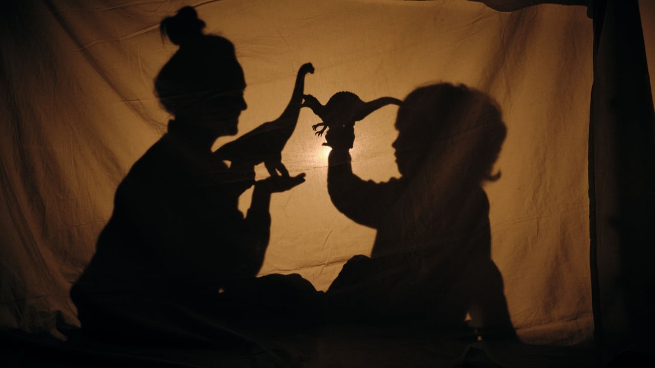 Side view of mother and son sitting behind curtain and playing with dinosaurs toys in shadow theatre