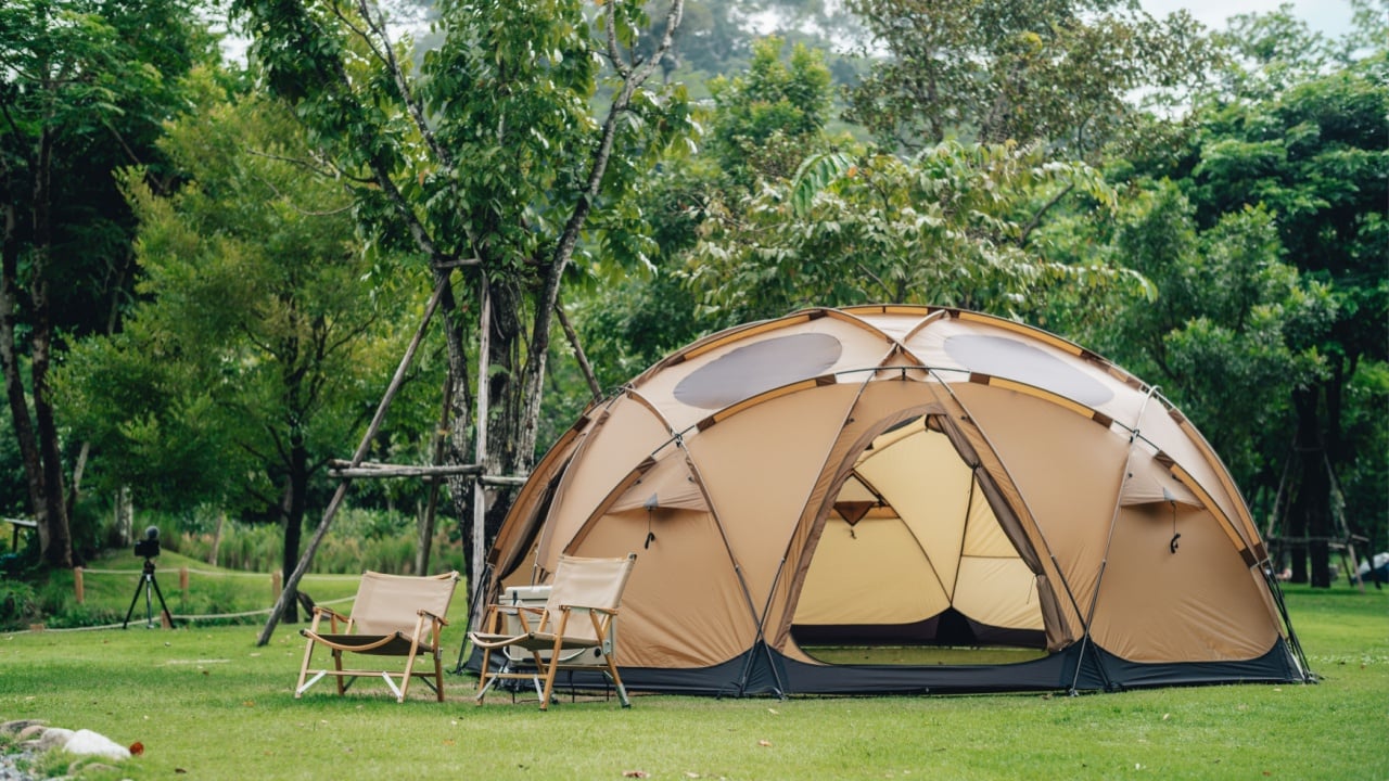 A big brown dome camping tent in a green grass with trees and mountains. Taken in an afternoon with a clear blue sky with white clouds. There're chairs, table and camping equipments nearby.