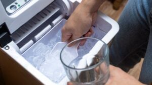 Closeup shot of a person's hand transferring bullet type ice cubes from a small portable ice maker into a clear glass with a small scoop.