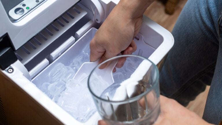 Closeup shot of a person's hand transferring bullet type ice cubes from a small portable ice maker into a clear glass with a small scoop.