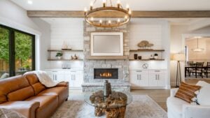 Living room interior with exposed wooden beams on the ceiling carmel colored leather sofa round wagon wheel chandelier fluffy white throw blanket hardwood floor view to kitchen and stone fireplace