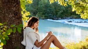 Young woman writing and drawing in notebook sitting under the tree near river on nature in sunlight. Closeness to nature inspires creativity. Urban escapism.