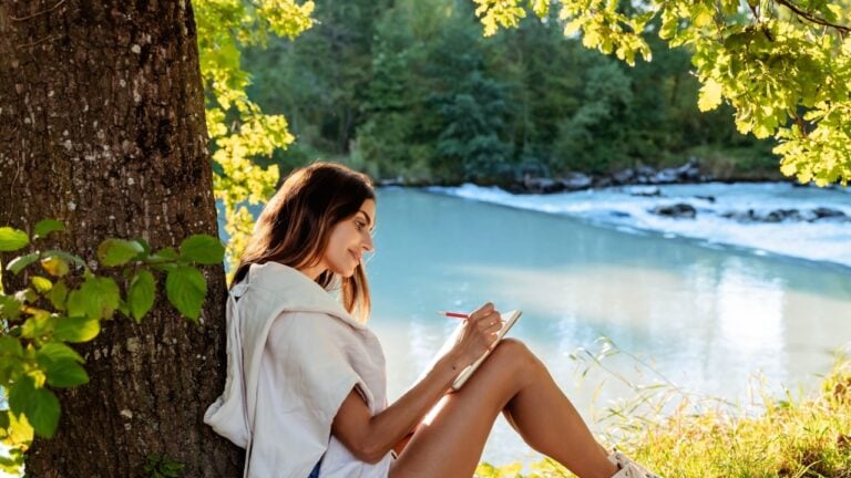 Young woman writing and drawing in notebook sitting under the tree near river on nature in sunlight. Closeness to nature inspires creativity. Urban escapism.