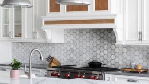 A kitchen detail with metal lights hanging above a large marble island, stainless faucet on a white farmhouse sink, and a marble hexagon tiled backsplash with white cabinets. No brands or labels.