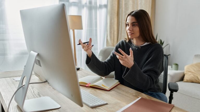 A young woman in a grey sweater creates engaging digital content from her home office desk, speaking passionately into her computer screen.