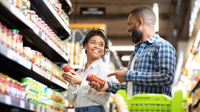 Happy African Family Couple Buying Food In Supermarket, Choosing Products Walking With Cart Along Aisles And Full Shelves Purchasing Groceries Together. Black Spouses Purchasing Essentials Together