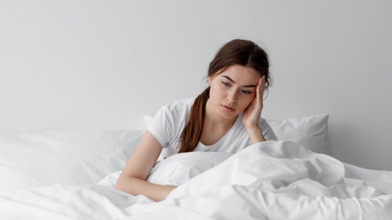 Sad european millennial female, worried, suffers from insomnia and headache. Lady sits on bed in bedroom at home, copy space. Migraines, health and mental problems, lack of sleep and depression