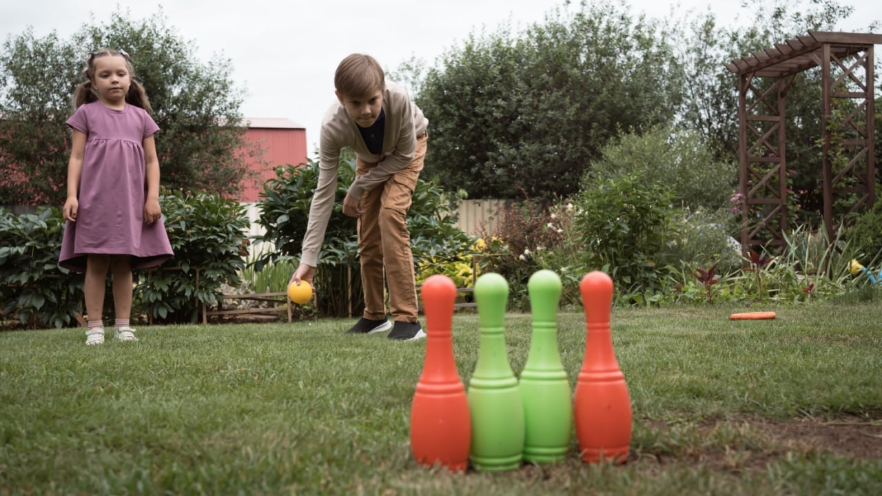 Two kids play toy bowling with colorful plastic skittles and ball on grass in garden or yard or park in summer day. Brother throws ball and little sister waits