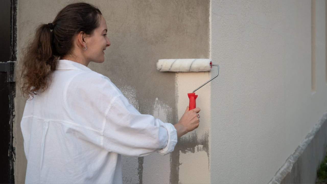 Painter is painting a house wall with a paint roller, applying a fresh coat of white paint on the building facade