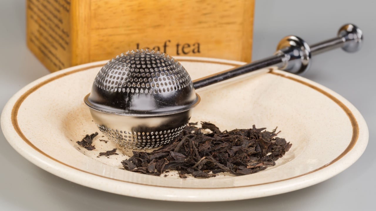 Closed spherical stainless steel tea strainer on a saucer with dried black tea leaves on a gray background, front view close-up in selective focus