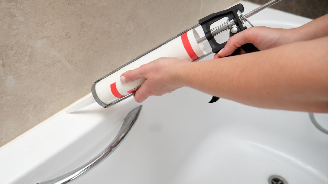 Hand applying sealant to bathtub corner using caulking gun. Home renovation and bathroom maintenance concept.