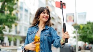 Young woman taking selfie with smartphone and selfie stick while walking down the street