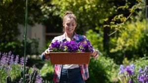 Woman is joyfully holding basket filled with vibrant purple flowers while standing in lush, sunny garden. The greenery surrounds her, enhancing the cheerful atmosphere.