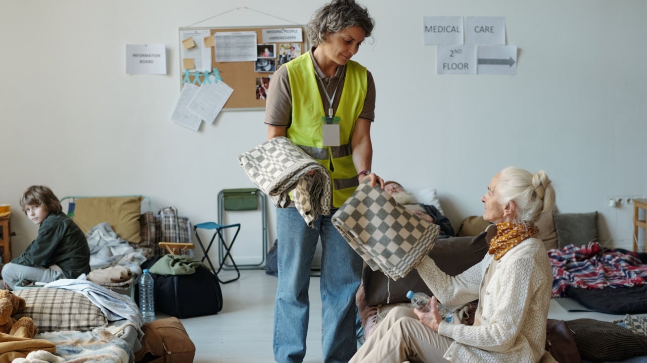 Smiling mature female volunteer in uniform passing folded blanket to senior woman sitting on sleeper in refugee camp