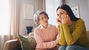 Two women sitting on couch in living room. Elderly female in pink sweater talking with daughter. Keeping hand on her shoulder while trying to support her. Girl looking upset and holding hands on chin.
