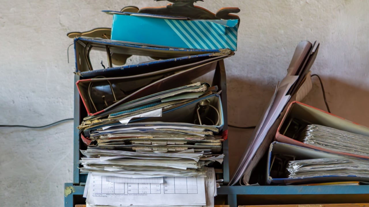 ruined shelf full of chaotically arranged binders, folders and documents. The furniture and papers look old and worn. In the background you can see a light cracked wall. an abandoned office.