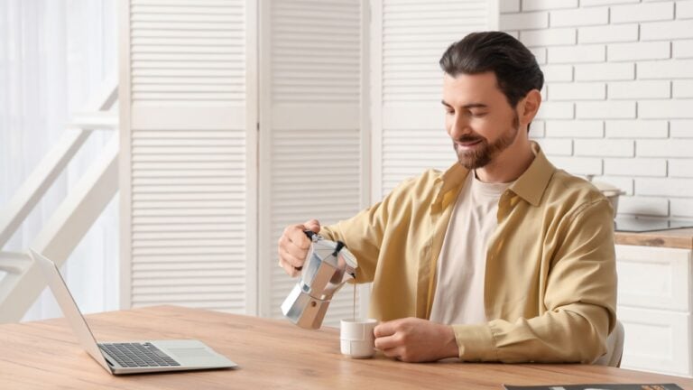 Young man with laptop drinking coffee in kitchen