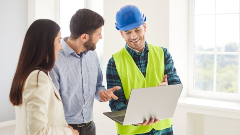 Happy couple husband and wife homeowners together with foreman, construction engineer, architect standing indoors, looking at digital house remodeling plan on laptop, discussing interior redesign
