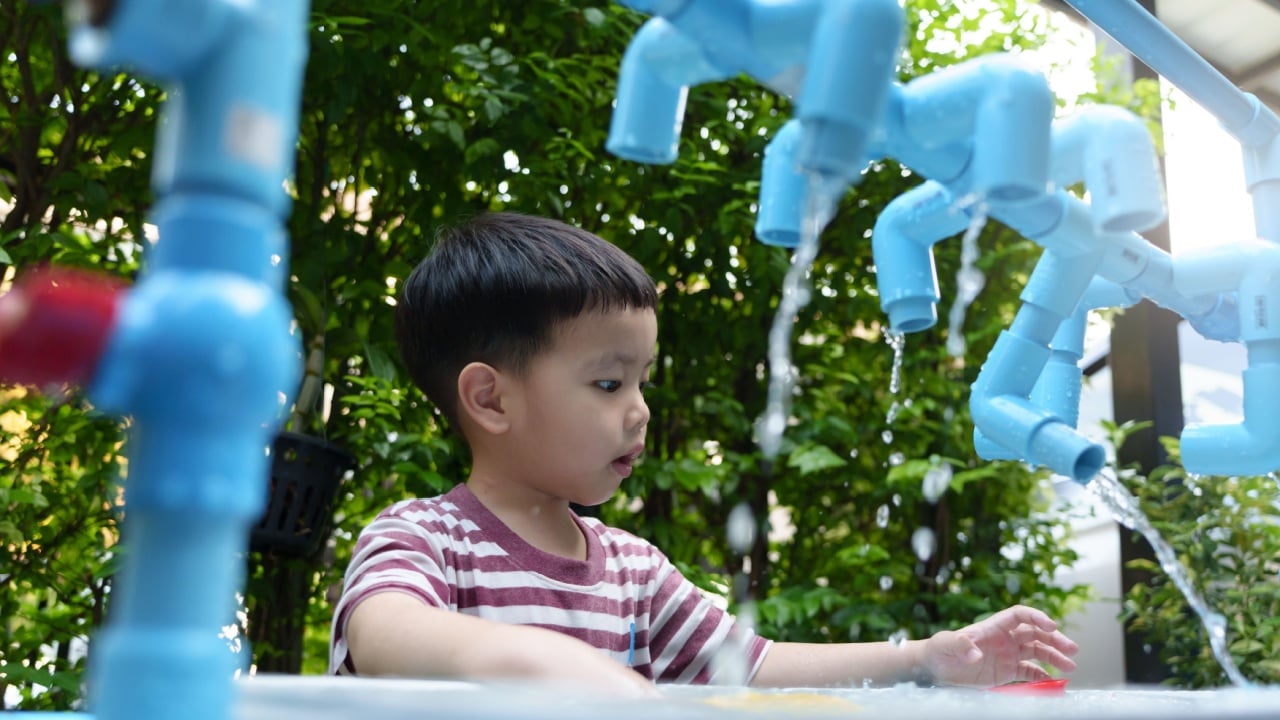 Asian boy engages in playful exploration with flowing water at a vibrant outdoor play area surrounded by greenery.