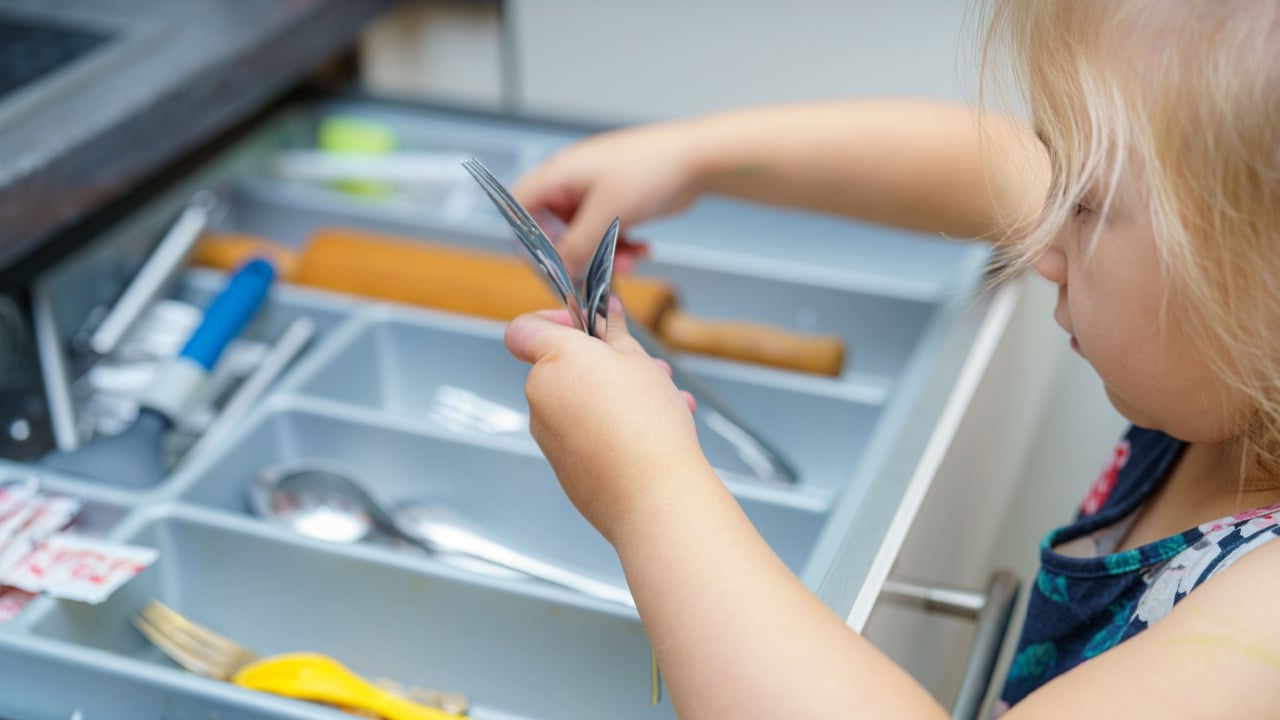 A young child is actively exploring a kitchen drawer filled with various utensils. The cheerful activity highlights curiosity and playfulness in a home environment during daylight hours.