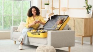 Young African-American woman with mobile phone packing suitcase for trip on sofa at home