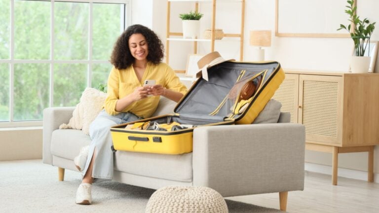 Young African-American woman with mobile phone packing suitcase for trip on sofa at home