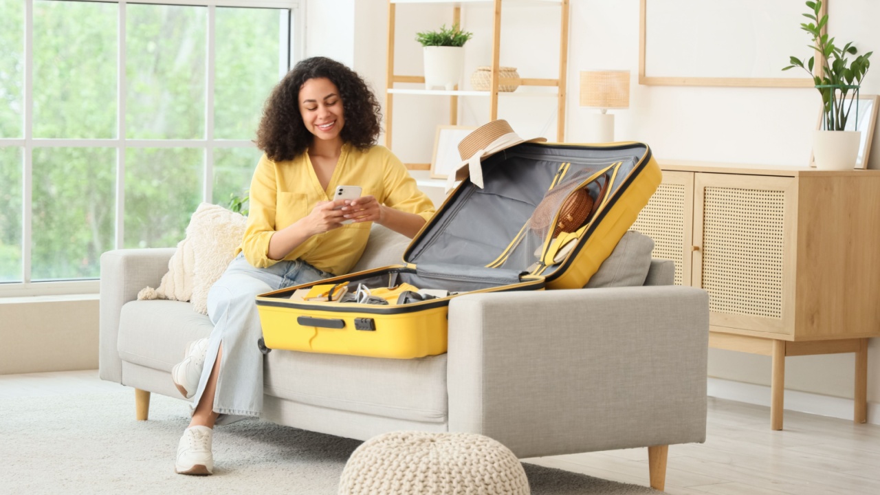 Young African-American woman with mobile phone packing suitcase for trip on sofa at home